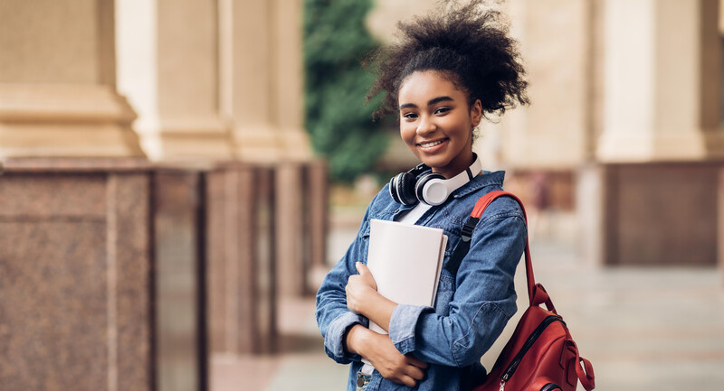 A high school student with a backpack and headphones stands outside a school building smiling happily 