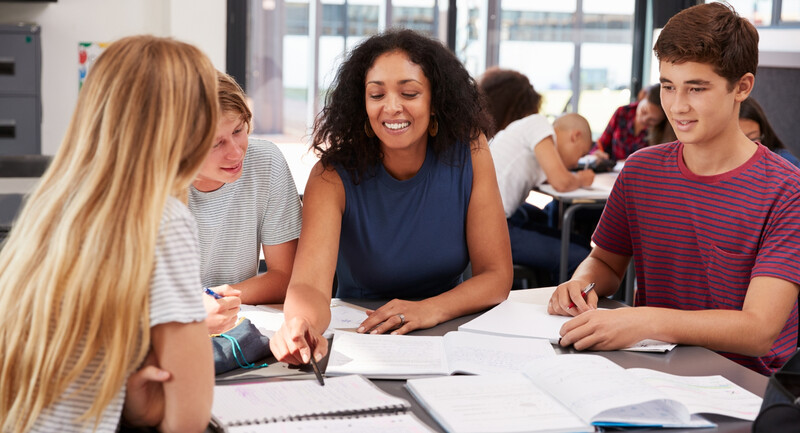 A new teacher interacts with a small group of students working on a project, smiling and pointing out something in a notebook.