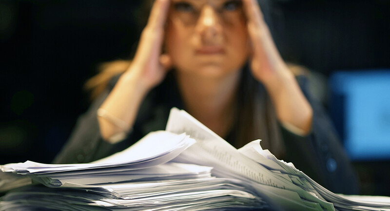 Large pile of papers stacked in front of teacher who appears frustrated and overwhelmed