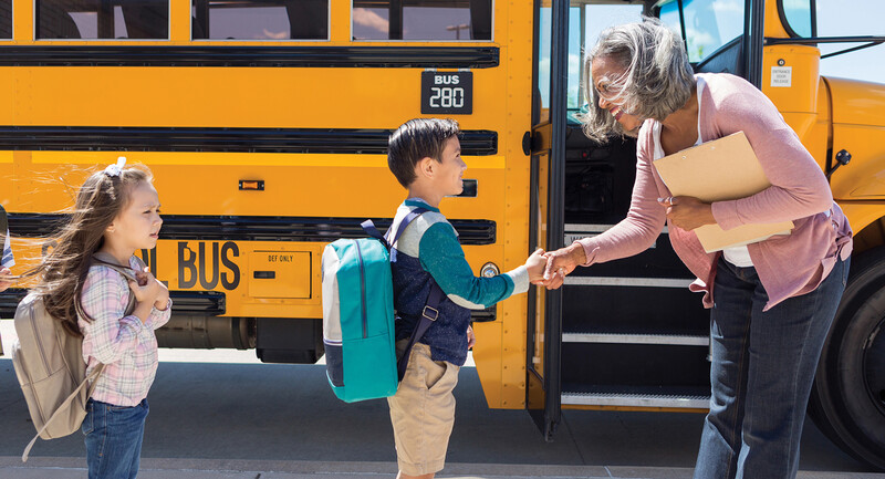 Photo of a teacher greeting young students in front of a school bus
